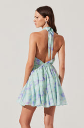 A woman is wearing a sleeveless dress with a pastel watercolor print in shades of green, purple, and blue. The dress features a halter neckline tied into a bow at the back of her neck and has a fitted waist with a flared, pleated skirt. The back of the dress is open, revealing her shoulders and upper back.